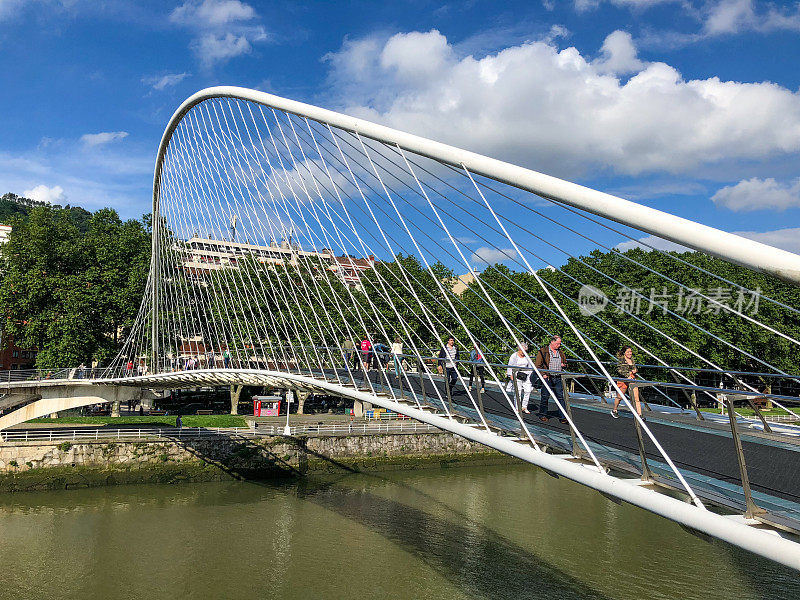 行人在未来主义的Campo Volantin人行桥上穿过Nervion河，桥上有一个系拱设计和弯曲的人行道